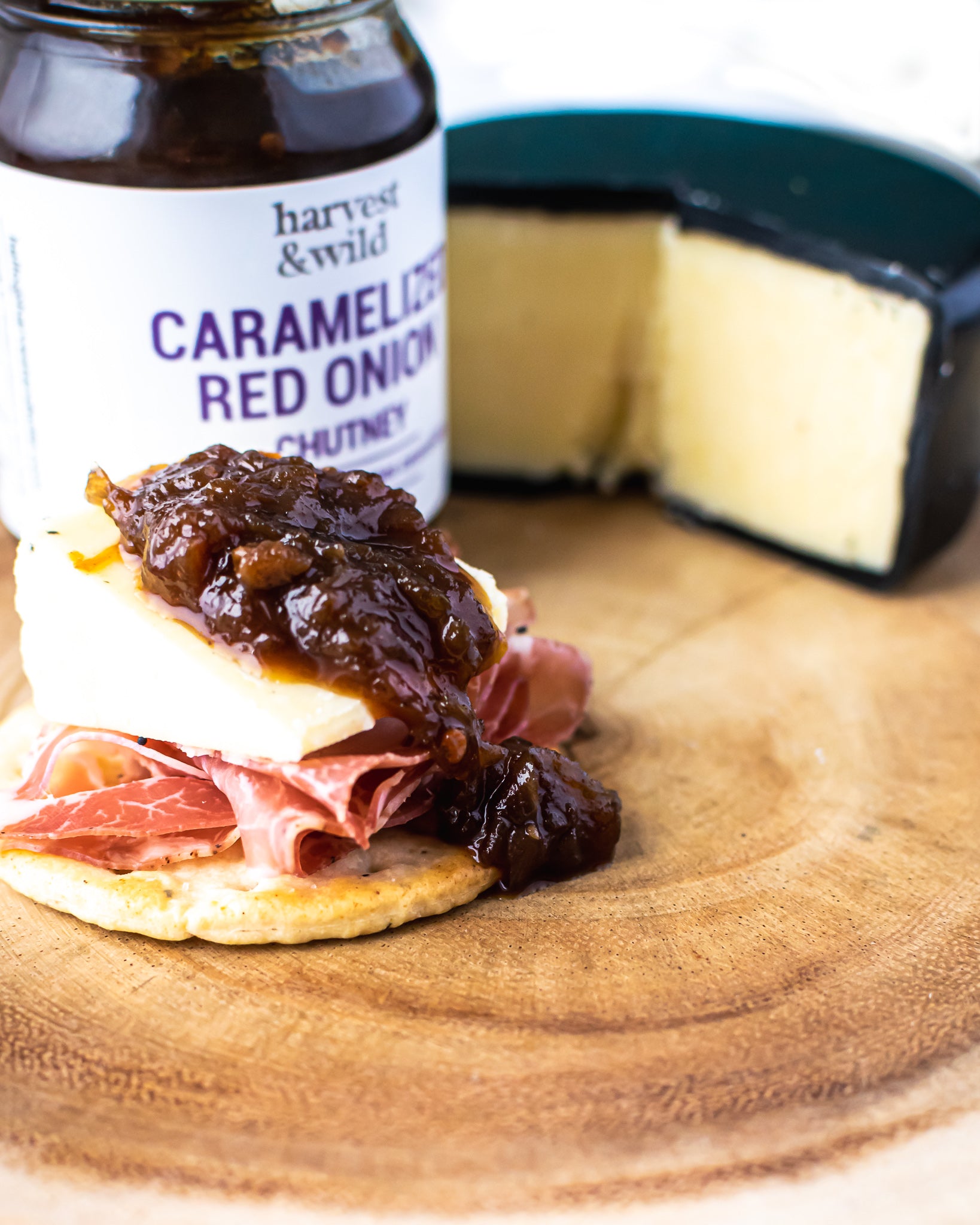 Caramelized Red Onion chutney on top of wedge of cheese and ham, wheel of cheese in in background - Harvest and Wild